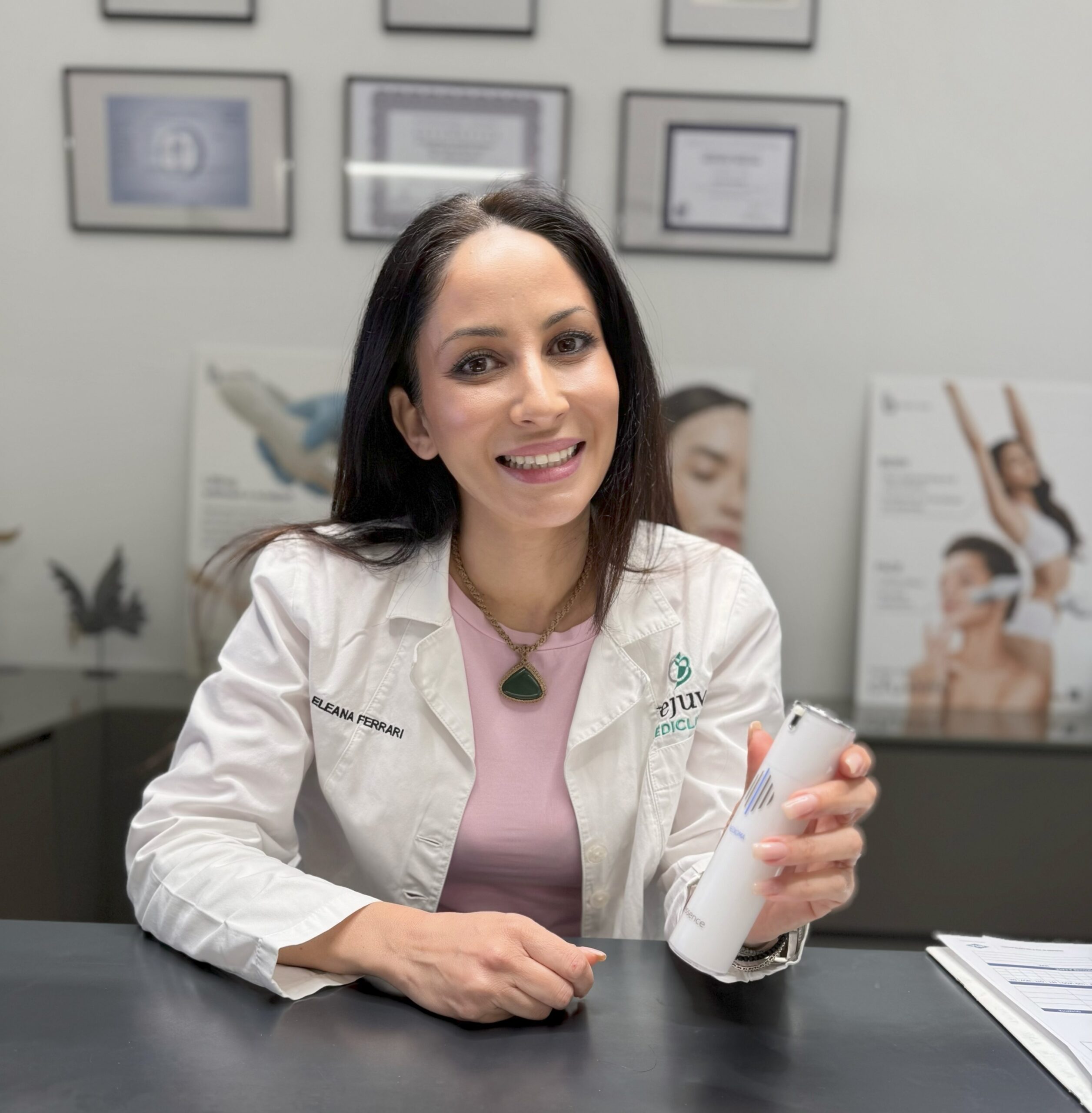 Woman in a medical coat with a name tag reading “Eleana Ferrari,” seated at a desk, smiling while holding a bottle of “Interactive” skincare; framed certificates and dermatology posters in the background.